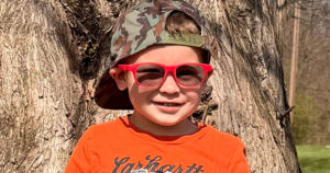 Wilder, wearing red prescription sunglasses, is sitting on a bench in the park and smiling for the camera.