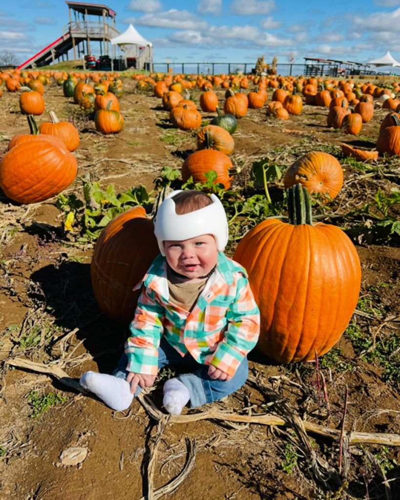 Wilder, wearing a cranial helmet, sits in a pumpkin patch.