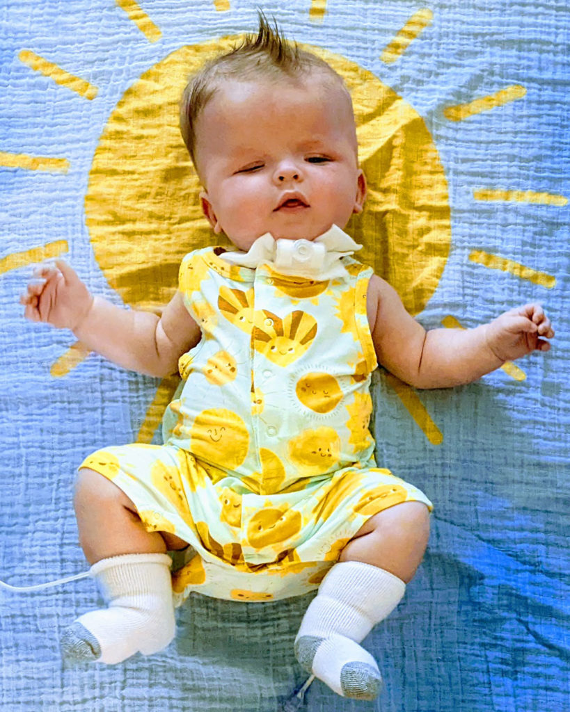 Sonny is wearing a onesie with smiling suns while lying on a blanket with a giant sun and blue sky background.