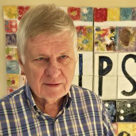 Man with a button up collared shirt and gray hair is standing in front of colorful and black and white tiles.