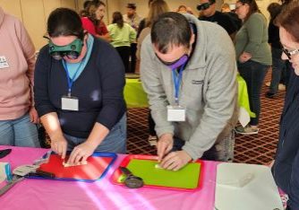 Two people are standing in front of cutting boards. One has a sleepshade on and the other has visual impairment simulator goggles on. They are both cutting food.