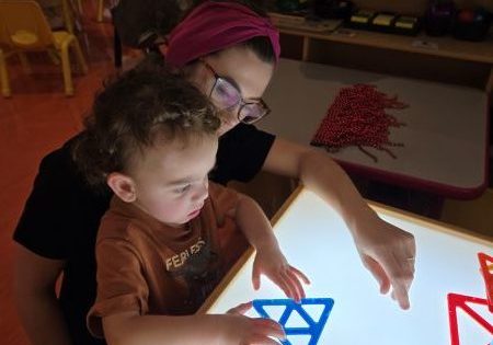 Child sitting with a teacher. The child has a large lightbox in front of them with a blue shape. The child is holding the shape.