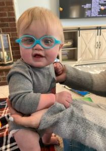 Baby is sitting up with the help of someone, has blue glasses and has a slight smile.