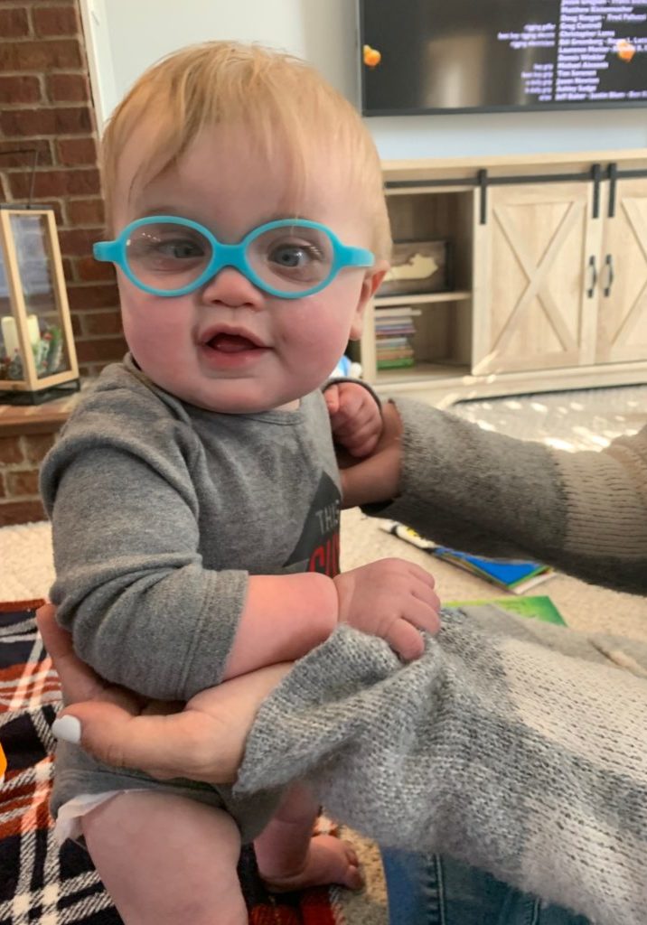 Baby is sitting up with the help of someone, has blue glasses and has a slight smile.