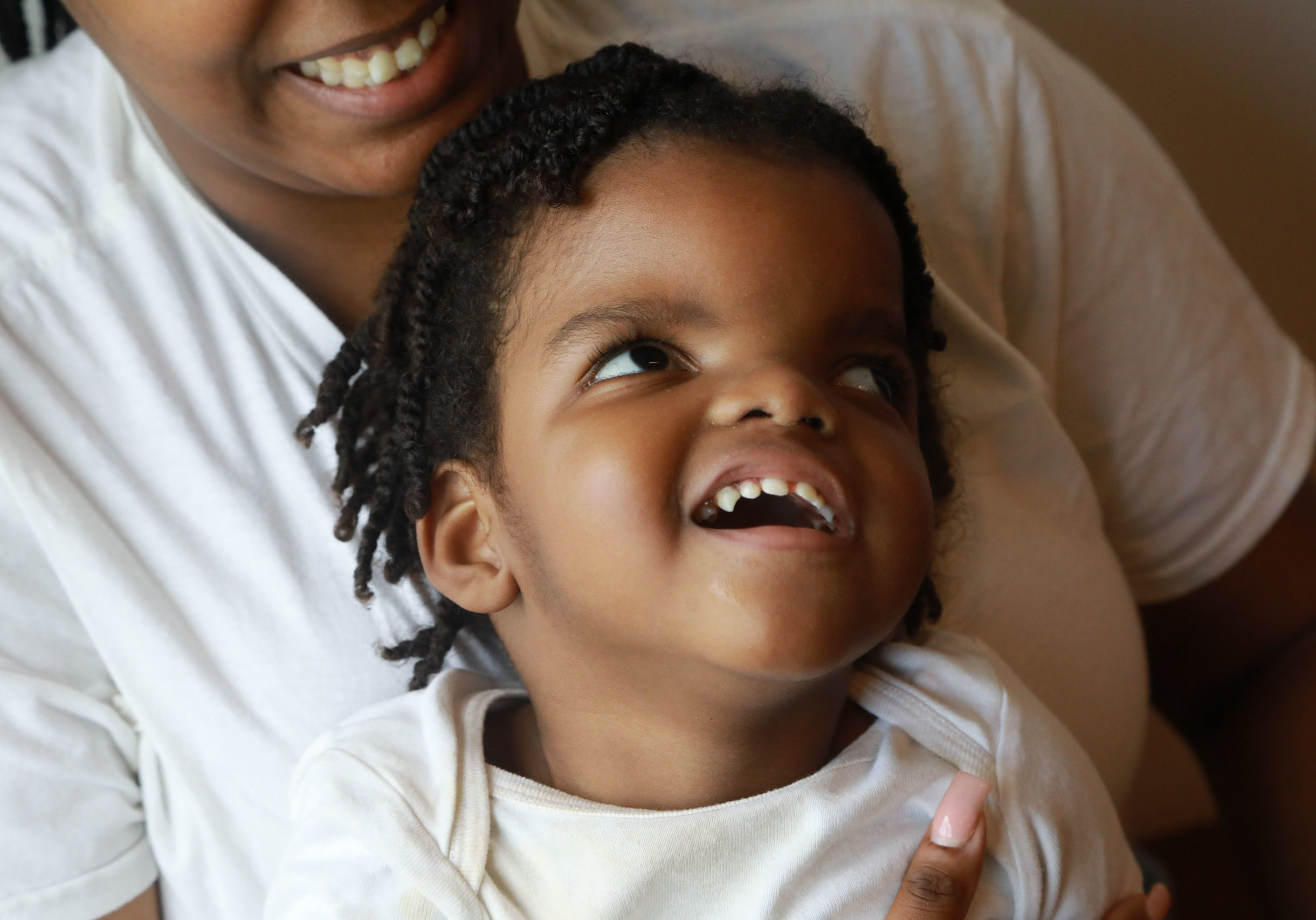 A boy is smiling while looking up and is sitting on the lap of someone who is also smiling. They are both in white shirts.