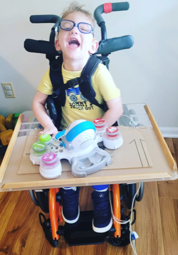 Noah_web Child smiling at camera while sitting in wheelchair with tray and toy.