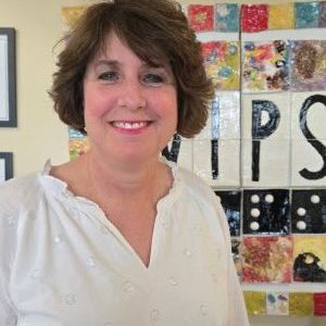 Woman in a white blouse and short dark hair is standing in front of artwork that says VIPS. She is smiling.