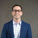 Man in a suit coat with a nice shirt and glasses on smiling at the camera.